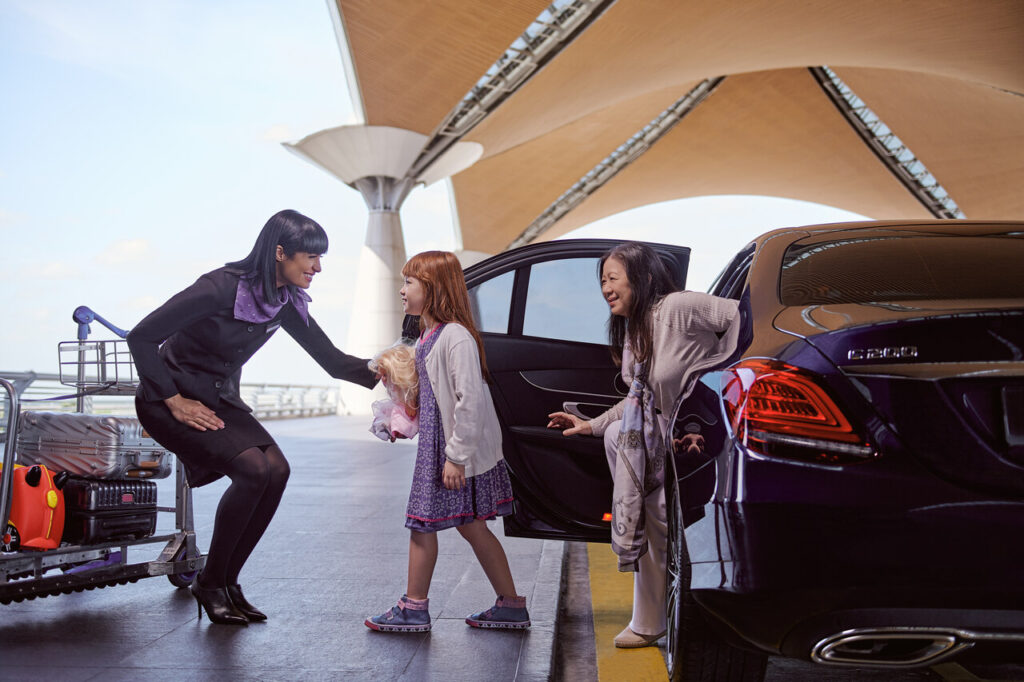 Using Beyond First by Plaza Premium, welcoming scene at an airport. A woman dressed in a professional uniform is bending down to greet a young girl who is holding a doll. The girl is standing next to a car, and another woman is seated inside the vehicle, reaching out towards the child. Luggage can be seen on a trolley nearby, and the setting is under a modern architectural structure, likely at an airport terminal. The atmosphere suggests a warm and friendly interaction, possibly part of a meet and greet service.