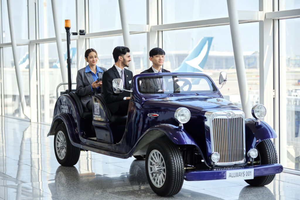 Using Beyond First by Plaza Premium Group, a vintage-style electric buggy used for concierge services at an airport. The vehicle is labeled "ALLWAYS 001" and is designed to transport passengers comfortably within the terminal. It features a classic car design with modern touches, suitable for indoor airport environments. The buggy is occupied by staff members dressed in formal attire, highlighting a service-focused approach to passenger assistance and convenience. The background reveals the airport's large windows and views of the tarmac, adding to the travel-themed context.