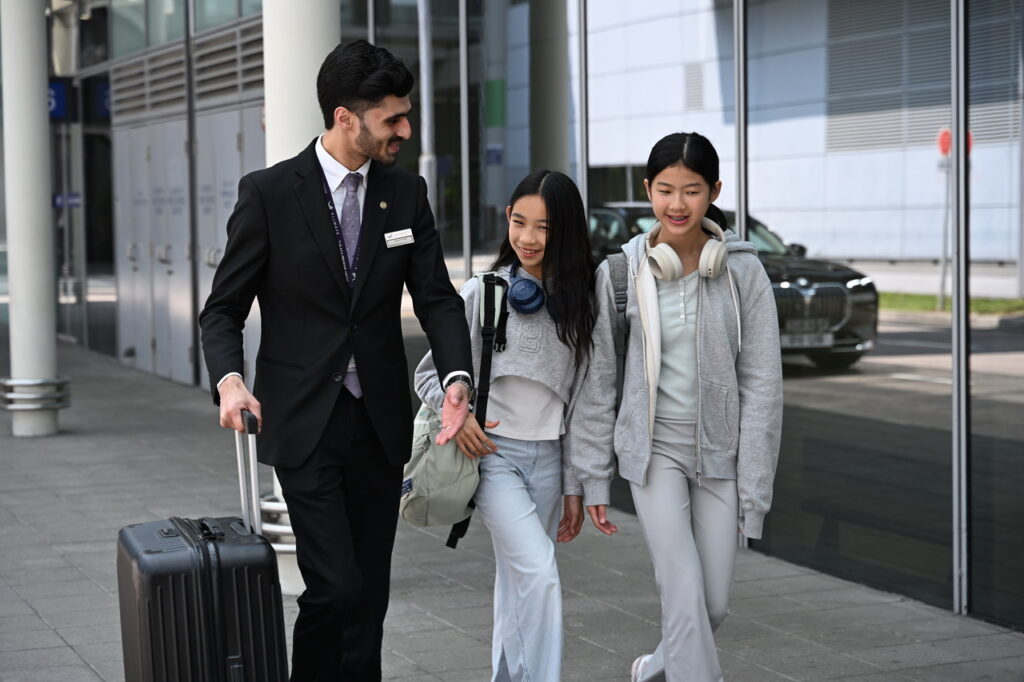Using Beyond First by Plaza Premium Group, A staff member, dressed in a formal suit with a name tag, is assisting two young travelers. The staff member is pulling a suitcase while engaging in conversation with the travelers, who are wearing comfortable travel attire and headphones. The background shows a modern building exterior, possibly an airport terminal, indicating a focus on customer service and travel assistance.