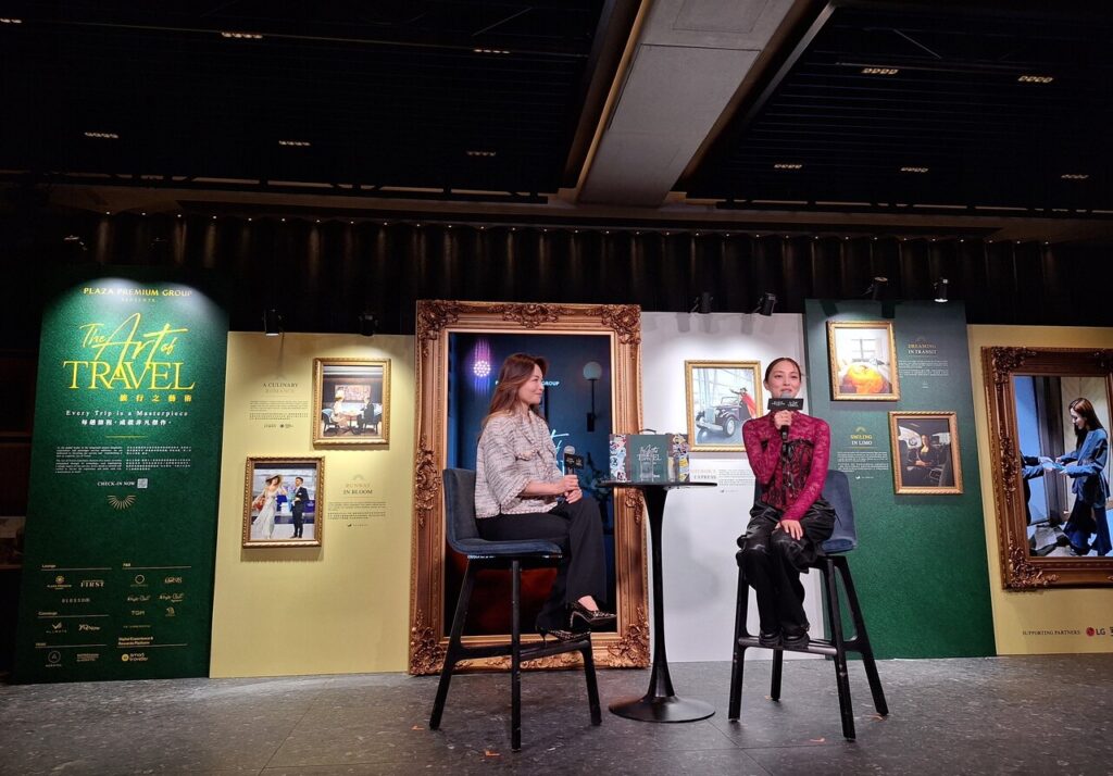 an interview or discussion event titled "The Art of Travel" hosted by Plaza Premium Group. The backdrop features elegant frames and promotional material, suggesting a focus on luxury travel experiences. Two individuals are seated on high stools, engaged in conversation. The setting is stylish and professional, with a well-lit stage and a sophisticated ambiance, indicating a formal presentation or interview related to travel and lifestyle.