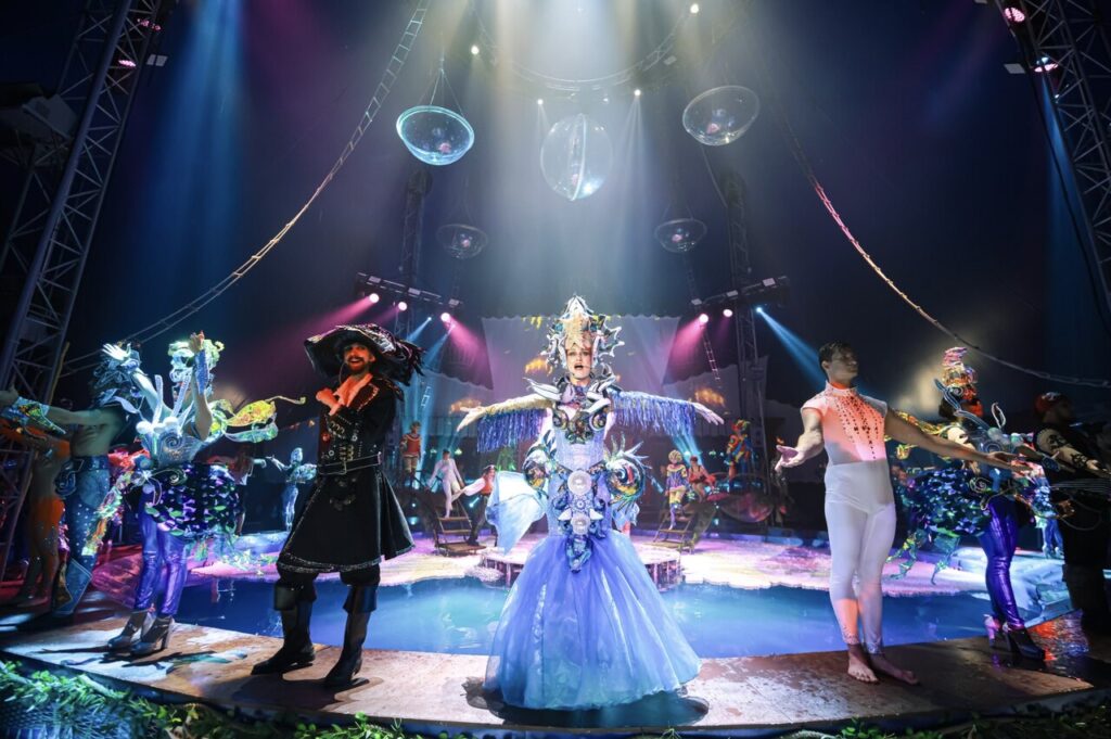 a dazzling stage performance filled with color and energy! The central performer stands out in a decorated blue dress with arms dramatically outstretched. Around them, other performers showcase a mix of costumes—one looking pirate-like, another in a sleek white bodysuit—adding to the theatrical feel. The stage is lit with dramatic lighting, and oversized reflective spheres hang from above, enhancing the atmosphere. The background features more performers and props, making the whole scene dynamic and immersive.