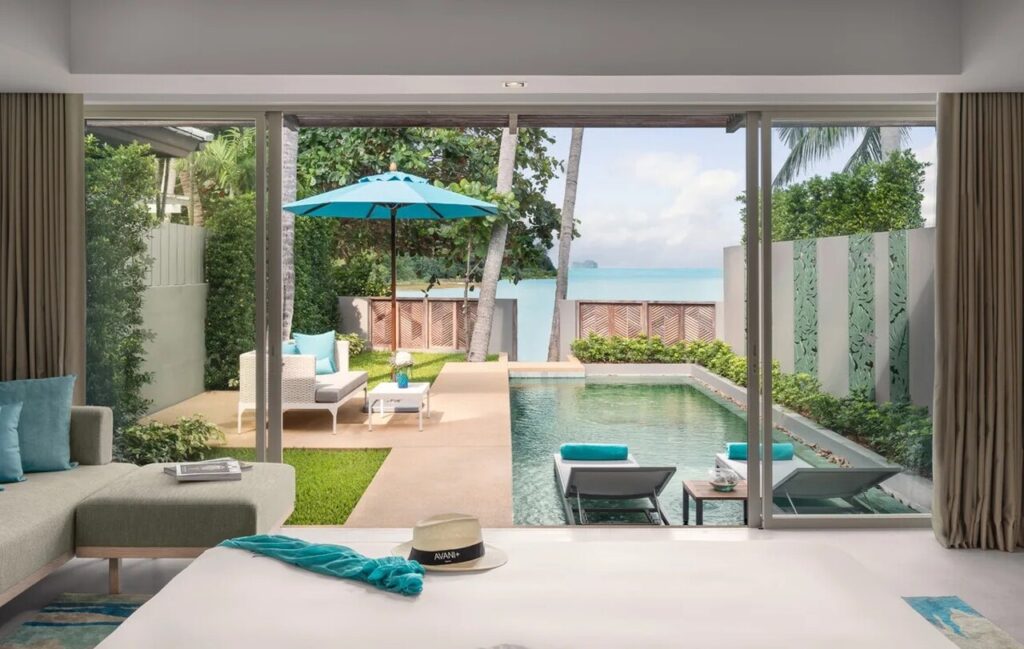 In Avani+ Samui, a stunning luxury outdoor retreat seamlessly connected to an indoor living space. The outdoor area features a private swimming pool surrounded by two elegant lounge chairs and a cozy patio setup, complete with a white wicker sofa and matching coffee table. A large blue umbrella offers shade over the seating arrangement, making the space feel inviting and relaxing. Lush greenery and palm trees frame the area, and the ocean view in the background adds to the serene atmosphere. Inside, there's a beautifully arranged bed with a turquoise throw and a stylish hat resting on it. A light gray sofa with turquoise pillows complements the airy and tranquil vibe of the space. Altogether, the scene suggests a luxurious resort or vacation rental designed for comfort and breathtaking views.
