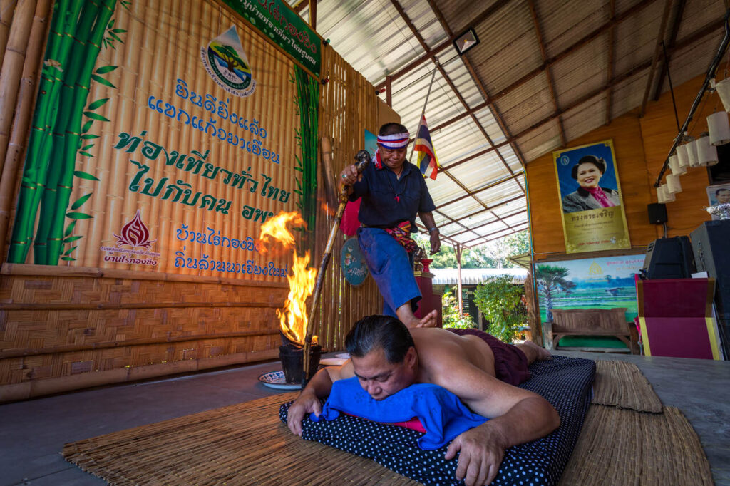 fascinating Thai cultural practice involving traditional healing or massage called Yam Khang. A person lies face down on a mat while another, dressed in traditional attire, stands on their back, holding a flaming staff near their body. The backdrop features a large banner with Thai text, bamboo decorations, a portrait, and a Thai flag hanging from the ceiling.