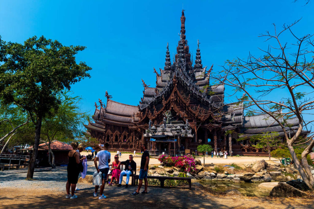 In Sanctuary of Truth, a stunning wooden temple with intricate carvings and multiple spires, exuding traditional Southeast Asian architectural elegance. The craftsmanship is breathtaking—every detail in the woodwork tells a story, making the structure feel both grand and deeply cultural. The central spire towers above, drawing the eye upward, while the surrounding trees and the bright blue sky create a beautiful contrast. In the foreground, flowers and rocks add a natural touch to the scene, framing the temple in a picturesque setting. The presence of people nearby gives the space a lively and welcoming atmosphere.