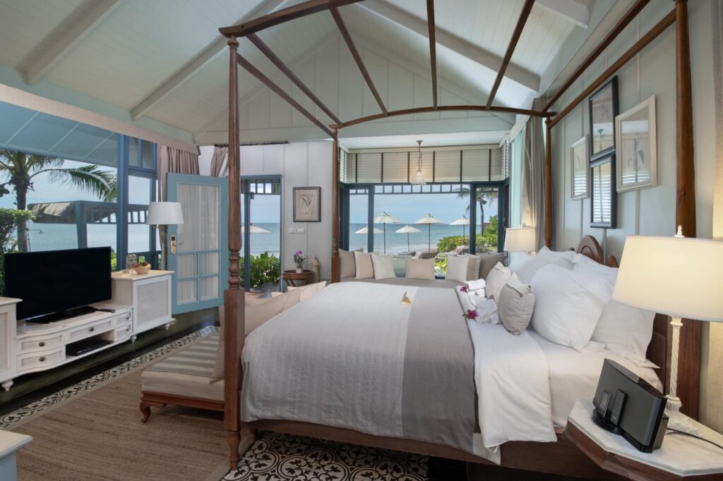In Devasom Hua Hin, This image showcases a stunning bedroom with a canopy bed overlooking the ocean. The room has a serene coastal style, featuring light colors and wooden accents. Large windows frame a beautiful view of palm trees, lounge chairs, and umbrellas on a patio, leading to the vast ocean beyond. The space feels like a high-end resort or vacation retreat, offering a luxurious, relaxing atmosphere.