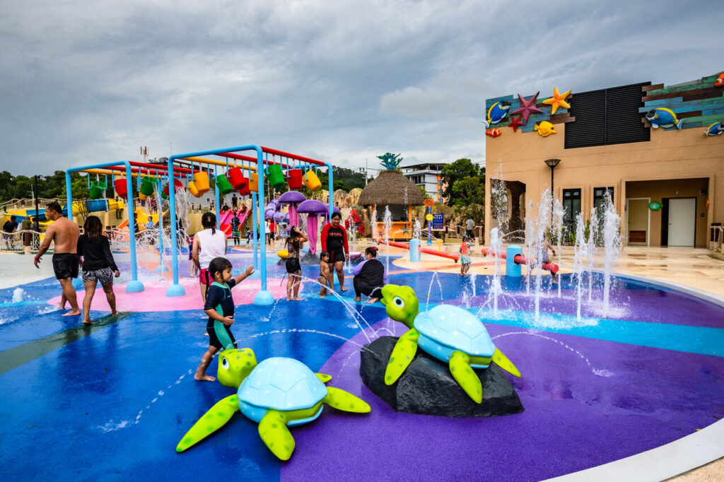 a lively water park scene filled with splashes and smiles! The park features two large turtle sculptures, each spraying water from their mouths, and several fountains shooting water upwards. There’s also a playful structure with hanging buckets that pour water once filled and jellyfish-shaped water features. In the background, a building adorned with aquatic-themed decorations—starfish and fish—adds to the underwater vibe. It looks like a fun-filled place where kids and families can enjoy water play and cool off on a sunny day.