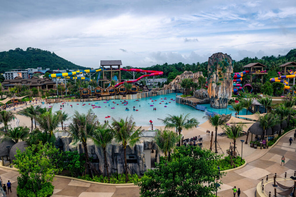 a vibrant water park filled with attractions. At the center, there’s a large pool with people floating on inflatable tubes, creating a fun and lively atmosphere. Surrounding the pool are palm trees and rocky formations, adding to the tropical aesthetic. In the background, there are towering water slides, including a red slide and a yellow-and-blue slide, promising excitement for thrill-seekers. Several buildings and structures are visible, including a tall tower that appears to be part of the slide complex. The park is enveloped by lush greenery and rolling hills, with some distant buildings peeking through. The cloudy sky suggests an overcast day, but the place still looks like an exciting destination for leisure and relaxation!