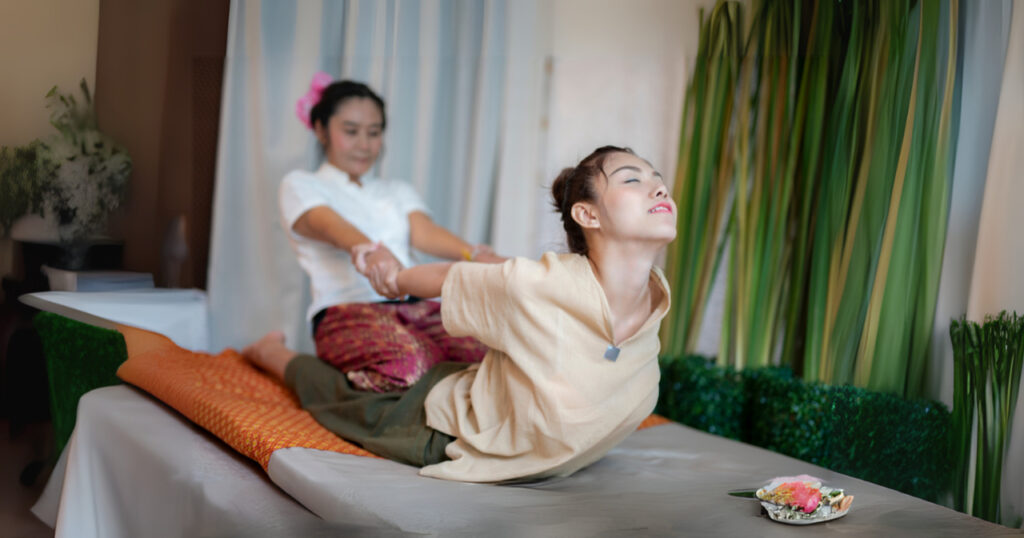 a person lying face down on a massage table while another person behind them provides assistance. The individual on the table is performing a stretch, arching their back and lifting their chest off the surface. The person assisting holds their arms and pulls them back gently.