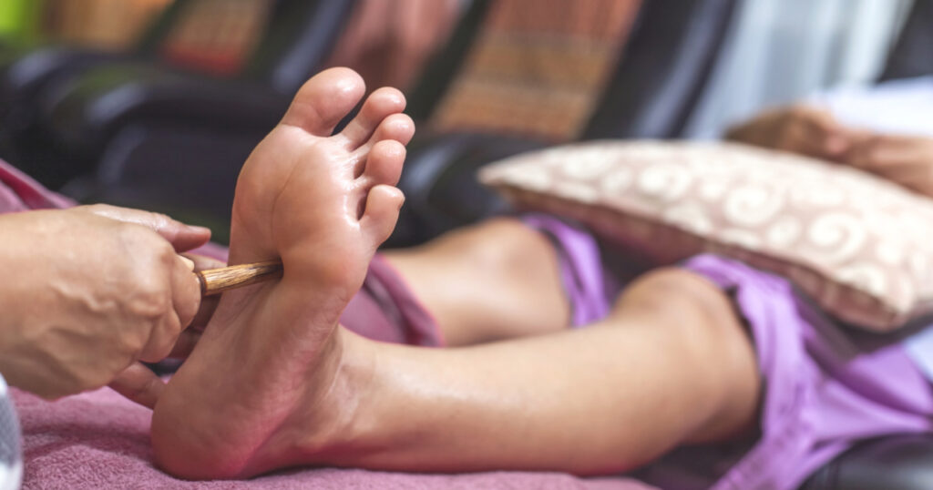 a close-up of a person receiving a foot massage. The individual is lying down with their legs extended, and another person is using a wooden tool to apply pressure to the sole of their foot. The background is slightly blurred, keeping the focus on the massage process. This depiction highlights a common relaxation and therapeutic practice, showcasing the use of specialized tools in foot massage techniques.