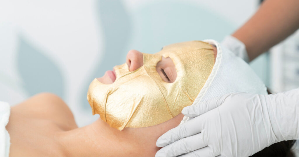 a person lying down with a towel or cloth wrapped around their head while receiving a facial treatment. A professional, wearing gloves, is gently touching the person’s face, possibly performing a skincare procedure. The setting appears to be a spa or clinical environment, emphasizing relaxation and professional care.