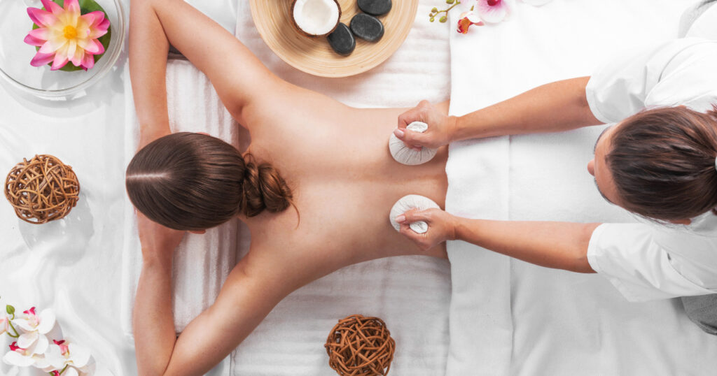 a person receiving a spa treatment. They are lying face down on a white towel with their hair tied up in a neat bun. A spa therapist is applying two round white objects to the person's back, likely part of a specialized therapy or relaxation technique.