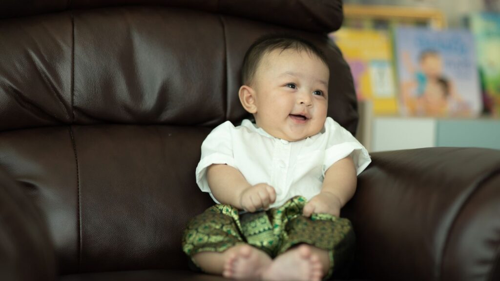 A cute baby boy sitting on a dark leather sofa, wearing a white shirt and patterned green shorts, with a softly blurred background for a warm, cozy feel.