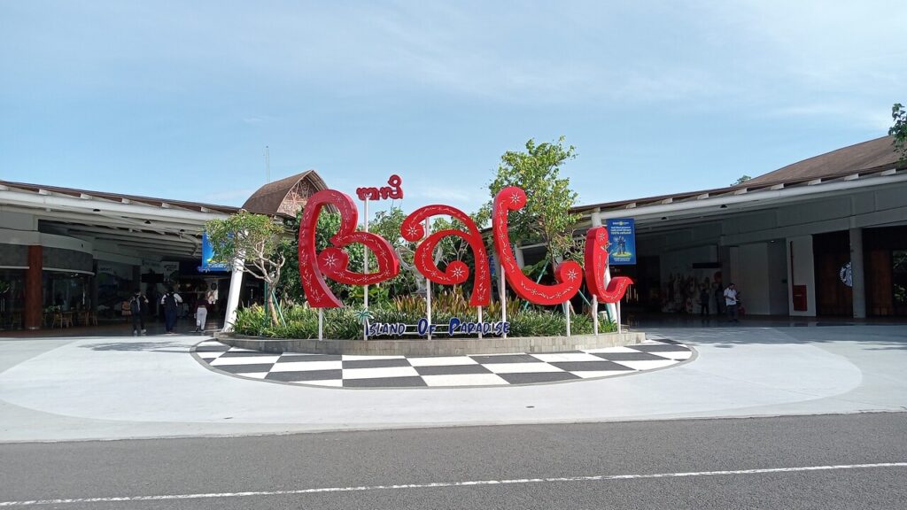 The “Bali” landmark sign welcomes travelers at Bali’s airport entrance, set in an open plaza under a bright blue sky.