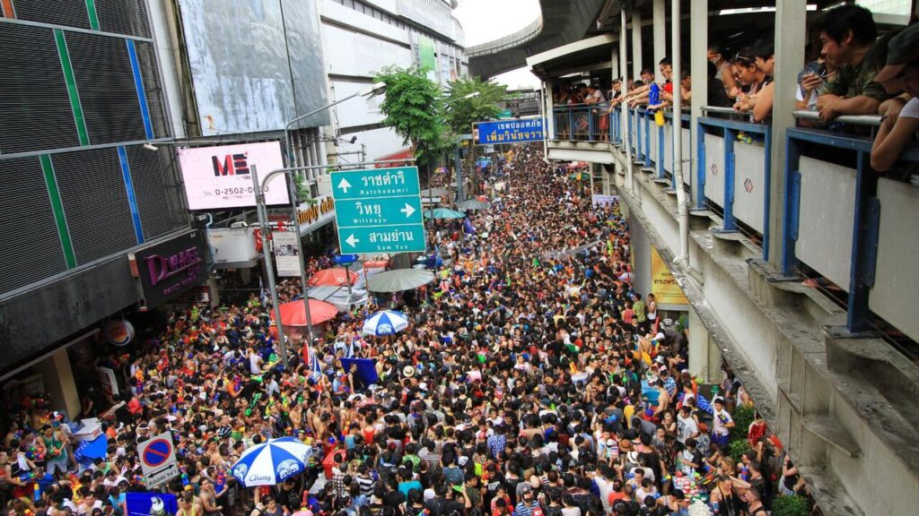 A massive crowd fills a busy Bangkok street during the Songkran, with people packed tightly beneath city signs and an elevated walkway lined with spectators.