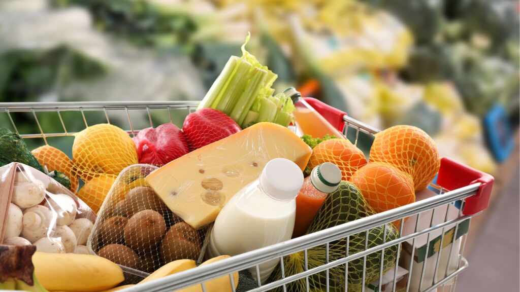 A grocery cart filled with fresh produce and staples, including oranges, avocados, mushrooms, cheese, milk, and vegetables in a supermarket setting.