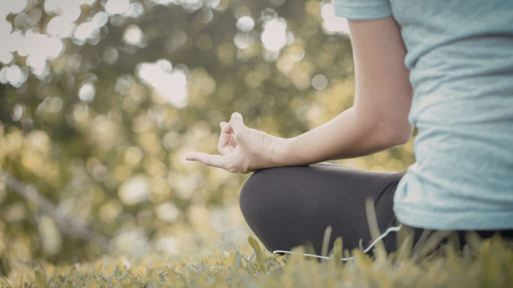 A person sits cross-legged on grass outdoors, practicing meditation with a relaxed hand mudra against a soft, sunlit background.