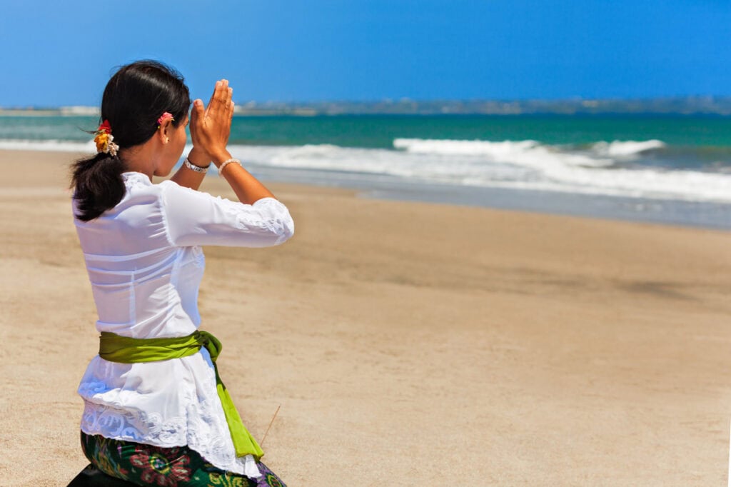A woman in traditional Balinese attire kneels on a sandy beach with hands pressed in prayer, facing the ocean under a clear blue sky.