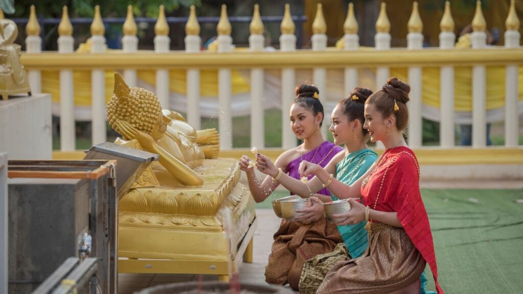 Women in traditional Thai dress kneel beside a golden reclining Buddha, gently sprinkling water in a serene temple blessing ritual.