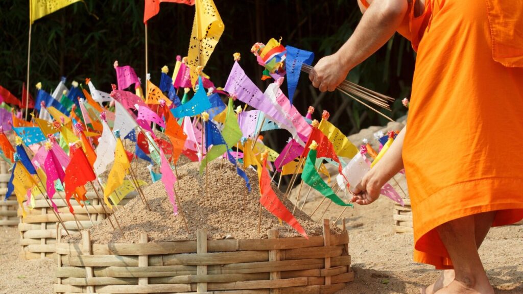 A Buddhist monk in orange robes places incense on a sand pagoda decorated with colourful flags, capturing a traditional Thai temple ritual outdoors. This is one of Thai traditional praying during Songkran.