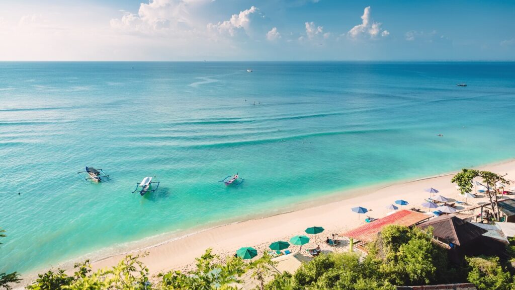 A tranquil tropical beach with clear turquoise water, small boats offshore, and colorful umbrellas lining a sandy shoreline under a bright sky.