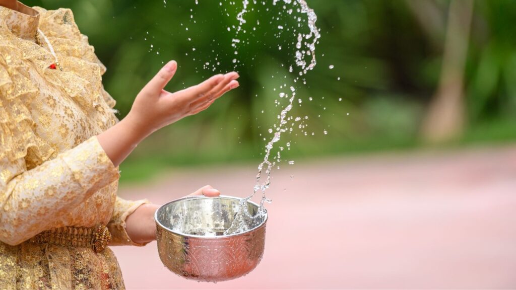 A lady in traditional attire holds a silver bowl as water splashes upward, capturing a serene cultural moment against a soft green outdoor backdrop. This is the traditional thai's blessing ceremony.