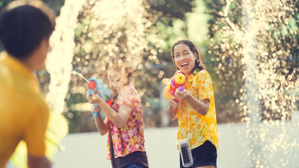 During Songkran, children in colorful floral shirts play a fun water-gun fight outdoors, surrounded by sparkling fountain spray in bright summer sunlight.